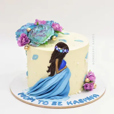 A beautiful "Mom to Be" cake featuring a hand-painted figurine of an expecting mother in a flowing blue dress, surrounded by delicate flowers.