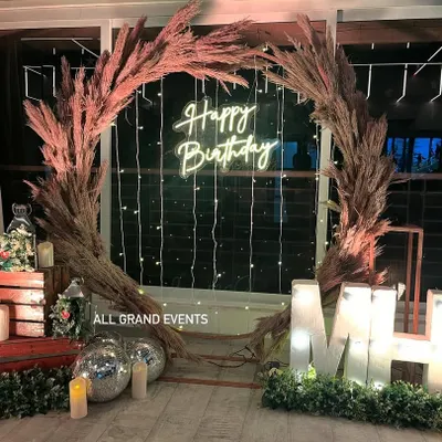 A beautiful rustic birthday backdrop featuring a large wreath made of pampas grass. The setup is personalized with light-up marquee initials and complemented by a neon sign and fairy lights.