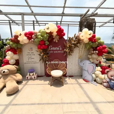 A beautiful 'Over the Moon' themed baby shower for Gauri. The setup includes two arched backdrops, a custom balloon garland in earthy red and green tones, and cute teddy bear and elephant props.