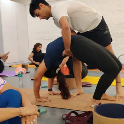 An instructor provides a hands on adjustment in Urdhva Dhanurasana, or Wheel Pose. This careful guidance helps the student lift and open their chest safely, ensuring the backbend is distributed evenly along the spine to prevent strain.