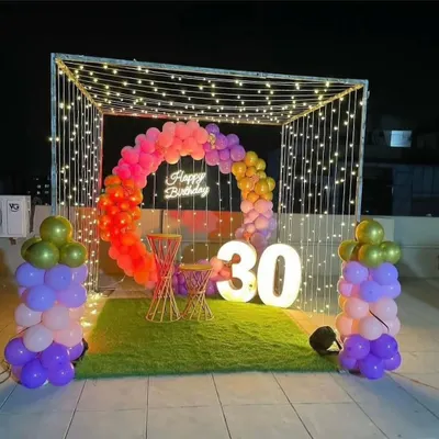 A vibrant 30th birthday setup on a terrace. This features a canopy of fairy lights, a colorful circular balloon garland, a 'Happy Birthday' neon sign, and large light-up numbers, all set on an artificial grass carpet.