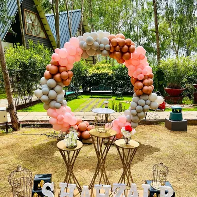 A beautiful daytime garden party featuring a circular balloon arch in muted tones of pink, grey, and bronze. The setup includes elegant dessert stands and personalized name lettering.