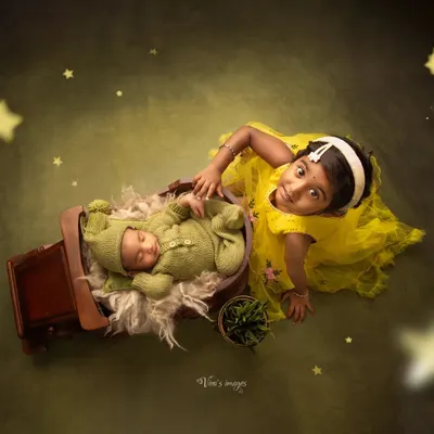 Big sister proudly watches over her sleeping newborn sibling. This creative shot uses a miniature wooden cart as a prop, adding a whimsical touch to this sibling and newborn portrait.