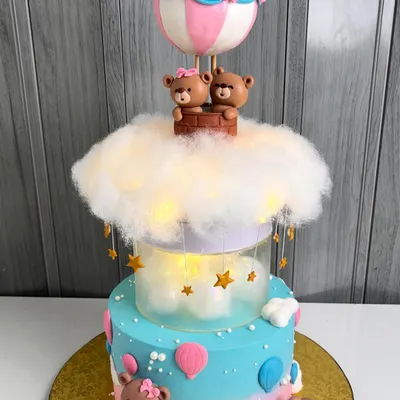 A magical two-tiered baby shower cake featuring teddy bears in a hot air balloon. The middle tier is illuminated with fairy lights behind a cloud of cotton, creating a dreamy, glowing effect.