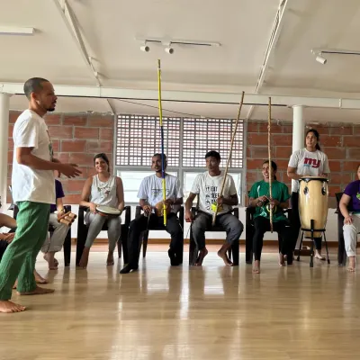 An instructor teaches the rhythm of the berimbau, the single-stringed instrument that leads the Roda. Participants learn to play instruments and contribute to the music.