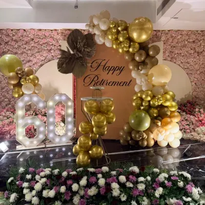 This was the main stage for a 60th birthday and retirement party. The setup featured a beautiful pink floral wall, a custom backdrop, marquee numbers, and a gold and white balloon arch, creating an elegant and celebratory focal point.