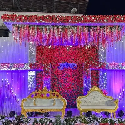 A grand reception stage featuring a massive red and white floral backdrop. The design incorporates hanging floral strings and ornate, royal-style seating to create a look of traditional grandeur.