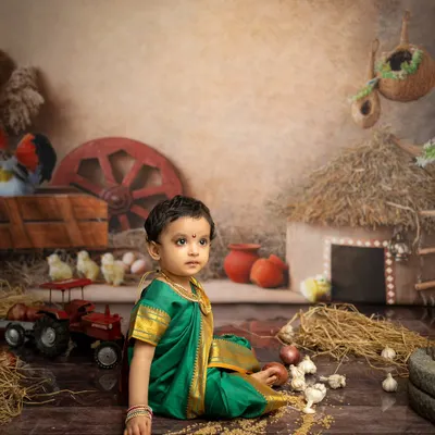 A thoughtful moment captured in our rustic Maharashtrian village set. The details, from the hay hut to the toy tractor, create an authentic backdrop for this traditional child photoshoot.