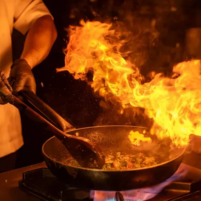 The raw energy of a live kitchen, captured as a chef creates a dish over high flames. This kind of dramatic action shot tells a story of skill, passion, and fresh, theatrical cooking.