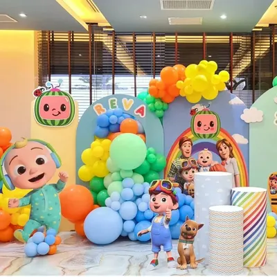 A bright and cheerful Cocomelon themed birthday party for Reva. This wide shot shows the full setup, including multiple character cutouts, themed plinths, and a rainbow of colorful balloons.