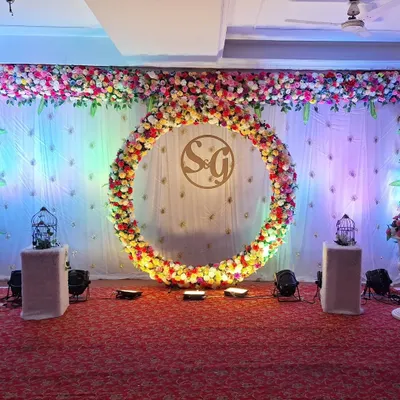 A classic and elegant engagement stage backdrop. The focal point is a large, circular ring completely covered in multi-colored flowers, with a custom monogram of the couple's initials placed in the center for a personal touch.