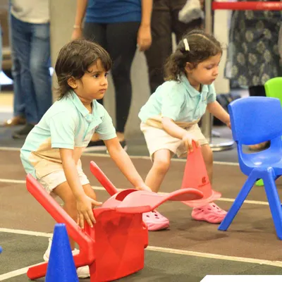 Giggles, races, and playful wins. Two children navigate an obstacle course with chairs, learning coordination and problem-solving during our fun-filled Sports Day.