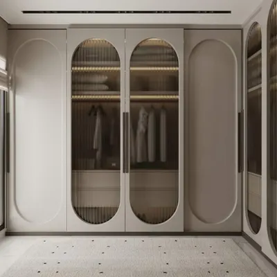 This walk-in wardrobe features custom floor-to-ceiling closets with elegant, capsule-shaped doors. The central doors have fluted glass panels and are backlit from within, turning the wardrobe into a stunning display piece while keeping clothes organized.
