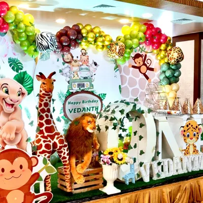 A close-up of a vibrant jungle safari theme for a first birthday. The setup includes a large "ONE" sign wrapped in vines, plush lions and giraffes, and a multi-colored organic balloon arch with zebra and leopard print accents.