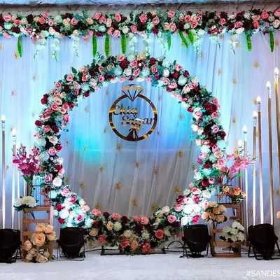 A stunning engagement stage centered around a large, lush floral ring made of pink and white roses. A custom sign with a ring icon hangs in the middle, all set against a beautifully lit white drape backdrop for a classic and grand celebration.