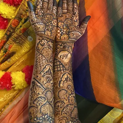 A complete bridal look featuring personalized initials within a heart on one arm. The design is dense with a mix of geometric grids, floral patterns, and traditional motifs, covering the arms fully up to the elbow.