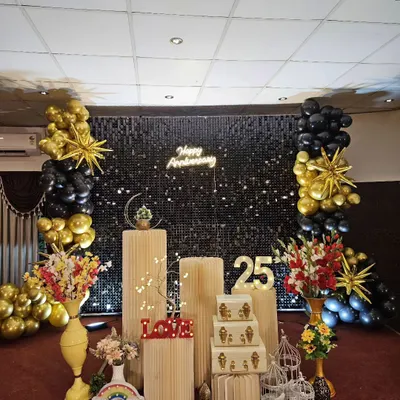 A sophisticated 25th anniversary decoration featuring a black shimmer wall and balloon columns in a chic black and gold color scheme. The setup includes a 'Happy Anniversary' neon sign and illuminated '25' marquee numbers.