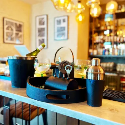 Elevate your home bar with the complete Bella Pelle Casa bar set. This image, set against a stylish bar backdrop, showcases the entire collection, including the tray, shaker, ice bucket, and toolset, all wrapped in sophisticated navy leather.