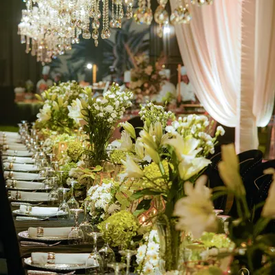 A beautiful dining setup under a canopy of crystal chandeliers, with long tables adorned with lush centerpieces of white and green florals, creating a classic and elegant atmosphere.