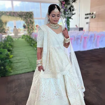 A breathtaking full-length shot of a bride in an all-white lehenga. The makeup is kept minimal and regal, with a clean bun adorned with velvet flowers, proving that elegance lies in simplicity.