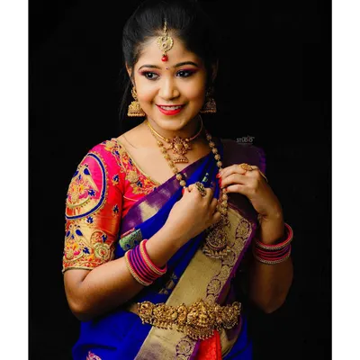 A classic South Indian bridal style from a model shoot, featuring a vibrant blue and pink Kanjeevaram saree. The makeup is bright and celebratory, with defined eyes and a cheerful lip color.