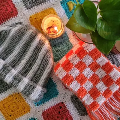 A cozy flat-lay of some of my crochet wearables against a multi-colored granny square blanket. This photo features a handmade striped beanie and a checkered scarf, showing the kind of stylish and functional items I create.