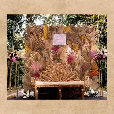 A cozy and stylish couple seating area with a boho theme. We used a beautiful peacock-style rattan bench set against a backdrop of dried and colored palm leaves, with a "Good Times" neon sign.