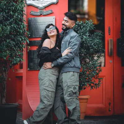 A fun and candid pre-wedding shot of a couple in casual, coordinated outfits in front of a vibrant red doorway at The Biere Club in Bangalore.