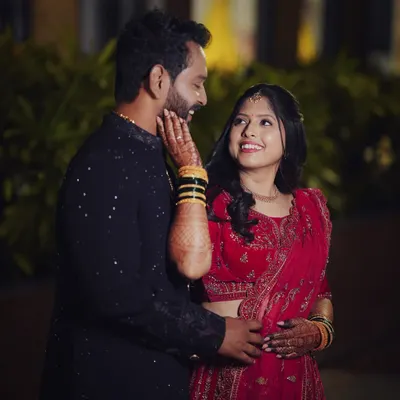 A candid and loving moment between a couple during their wedding festivities. The bride, in a vibrant red lehenga, looks up at her partner with a bright smile as he gently holds her face.