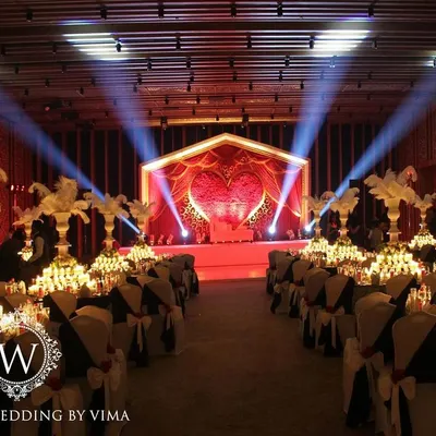 A glamorous anniversary celebration with a dramatic red and black theme. The room is filled with candlelit tables, large white feather centerpieces, and a stunning stage with a heart-shaped light installation.