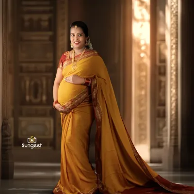 A full-length portrait showcasing the beautiful drape of a yellow and red silk saree. The architectural backdrop and soft lighting add a touch of grandeur to this traditional maternity photo.