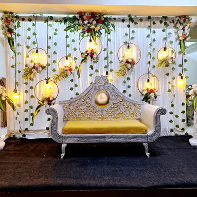 This reception stage combines rustic and industrial elements, featuring hanging floral hoops with exposed Edison bulbs. The design is set against a backdrop of green vines and white drapes, creating a warm, romantic glow.