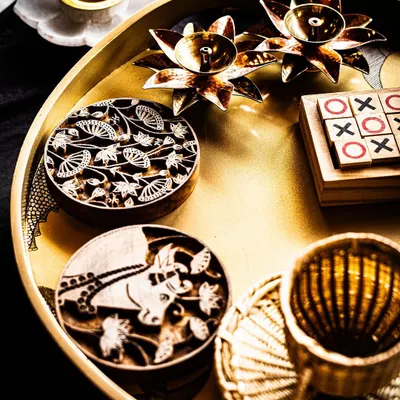 A curated tray of artisanal gifts, showcasing the beauty of traditional craftsmanship. It features hand-carved wooden printing blocks, brass lotus candle holders, and a wooden tic-tac-toe game, perfect for a memorable guest welcome hamper.