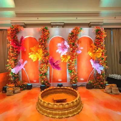 For this Haldi, I designed a unique stage with an orange backdrop and oversized, colorful paper flowers. The traditional brass urli in the center is ready for the ceremony, blending modern art with sacred ritual.