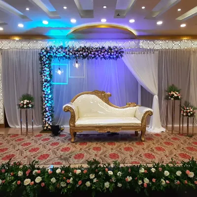 A sophisticated stage for an engagement ceremony. The decor features an L-shaped floral arrangement, elegant white drapery, and modern lighting fixtures, with a classic white and gold chaise lounge.