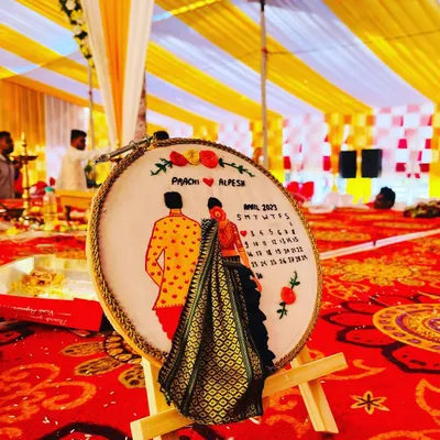A close-up look at a custom wedding hoop. This piece features detailed thread-work portraits, the couple's names, and a calendar marking their wedding date. The highlight is the bride's miniature saree, created with real fabric for a unique 3D effect.