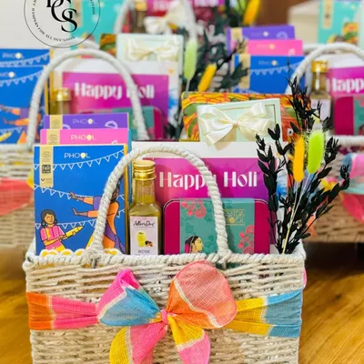A vibrant Holi gift hamper in a woven basket. It's filled with festive essentials like organic colors from Phool, a bottle of rose sherbet, and other treats, all tied with a colorful tie-dye ribbon.