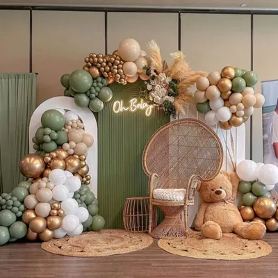 A stunning boho-chic baby shower with an olive green, white, and gold color scheme. This setup includes a wicker peacock chair, pampas grass, jute rugs, and an "Oh Baby" neon sign for a trendy and elegant look.
