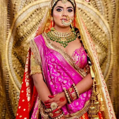A regal bridal look featuring a multi-toned lehenga with pink and orange hues. The makeup is focused on bold eyes and is complemented by heavy, traditional jewelry.