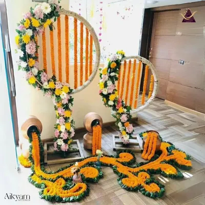 A creative and artistic floral installation for a home event. This decor features marigold garlands flowing from traditional pots, set against circular frames wrapped in flowers, creating a modern yet traditional look.