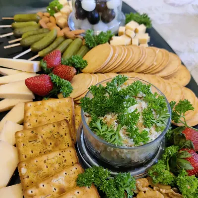 A beautiful cheese platter designed for a gathering. It features a variety of hard and soft cheeses, our homemade crackers, pickled gherkins, fresh strawberries, nuts, and a savory dip garnished with parsley.