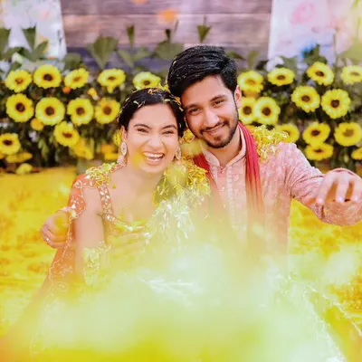 A fun and playful Haldi shot, with the couple smiling for the camera amidst a cloud of yellow turmeric powder.