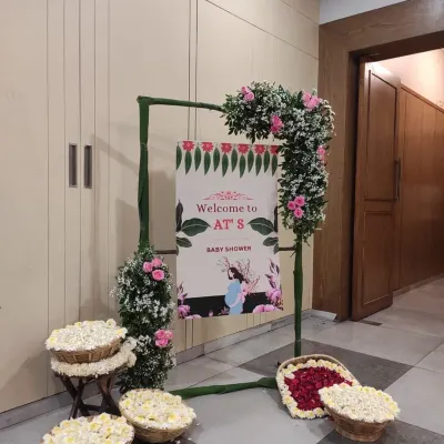 A simple and elegant welcome setup for a traditional baby shower. The sign is framed with bamboo and decorated with pink roses and fresh greenery, while baskets of white flowers (mogra) add a fragrant, traditional touch.