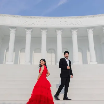 A dramatic, wide-angle shot on the steps of a grand, white library set. The bride's red gown creates a stunning visual pop, perfect for a movie-poster style pre-wedding photo.