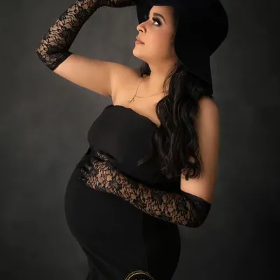 A chic and classic maternity portrait. The mother-to-be wears a simple black dress, elegant lace gloves, and a stylish hat for a timeless, fashionable look.