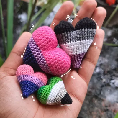 A handful of my handmade crochet pride hearts. This photo shows the bisexual, asexual, and aromantic pride flags, available in different sizes for pins, keychains, or earrings. This is queer art you can wear.