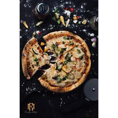 A top-down shot of a vegetable pizza, with a slice pulled away to show the cheese stretch. The dark, messy-on-purpose styling with scattered flour and toppings gives it an authentic, handcrafted feel.