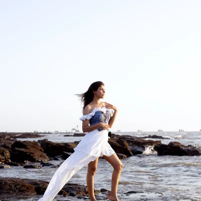 This shot captures a dramatic, windswept look on a rocky beach. The full-length view showcases the unique silhouette of the custom-designed dress with its flowing train, creating a powerful image for a fashion portfolio.