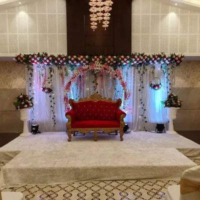A beautiful reception stage setup in a modern banquet hall. The floral arch, soft drapery, and a classic red velvet sofa create a rich and romantic focal point for the couple.