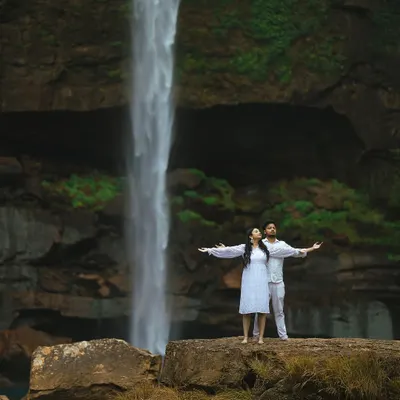 A couple stands on a rock ledge, arms outstretched, embracing the power of the waterfall behind them. This wide shot captures the scale of the landscape and the feeling of freedom.